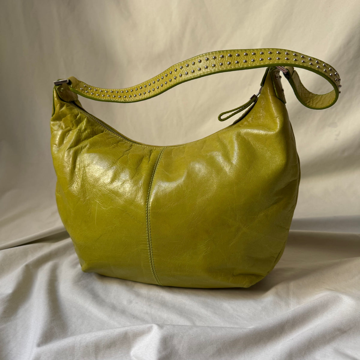 A yellow-green leather hobo shoulder bag with a matching studded strap, displayed on a draped white fabric background.