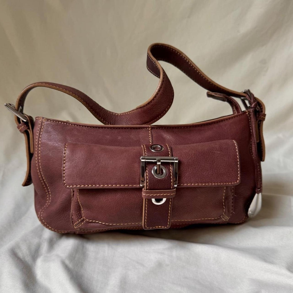 A brown pink rose coloured  leather shoulder bag with a front flap and silver buckle closure, resting on a draped white fabric background.