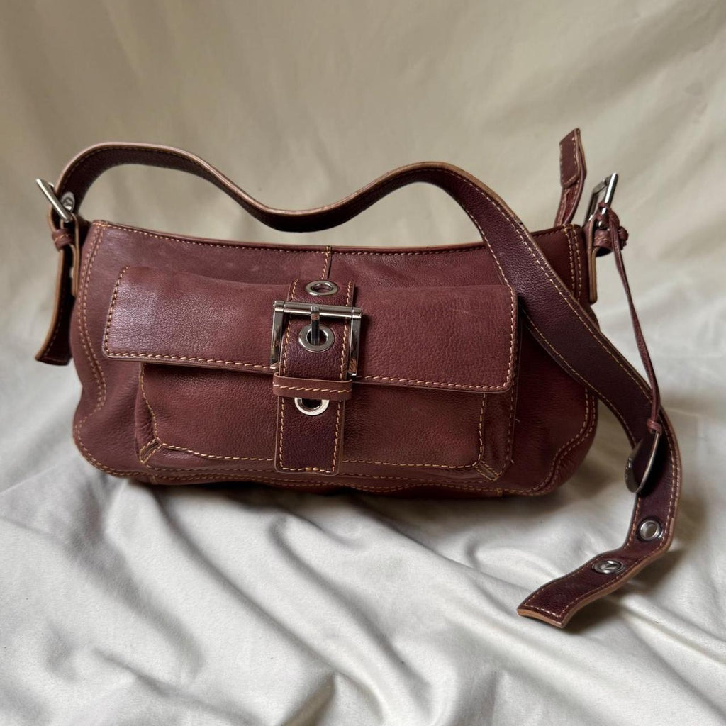A dark reddish-brown leather shoulder bag with a front flap and silver-tone buckle, resting on a draped cream satin fabric background.