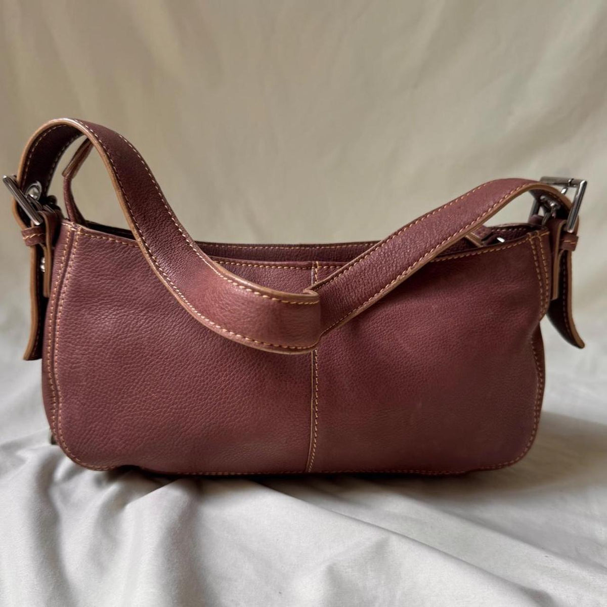 A reddish-brown Y2K vintage leather shoulder bag with a wide top handle and silver-tone hardware, photographed against draped light fabric.
