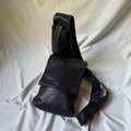 A black leather crossbody bag with a wide strap and a front flap pocket, photographed on a draped off-white fabric background.